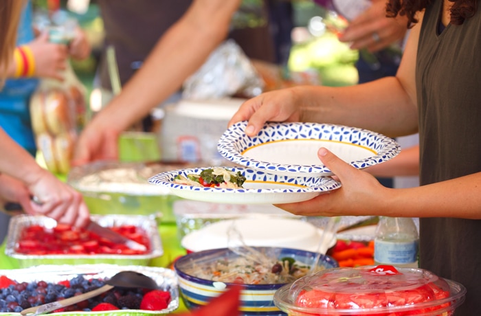 woman holding two plates, adding foor to the plates with potluck style table in the background signifying a back to school block party