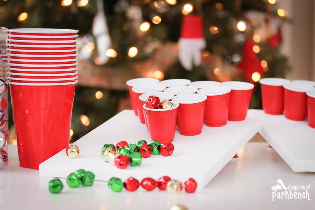 2 sets of 10 Miniature red plastic cups lined up in a triangle to make a jingle bell toss game, with Christmas colored jingle bells shown in front of the cups