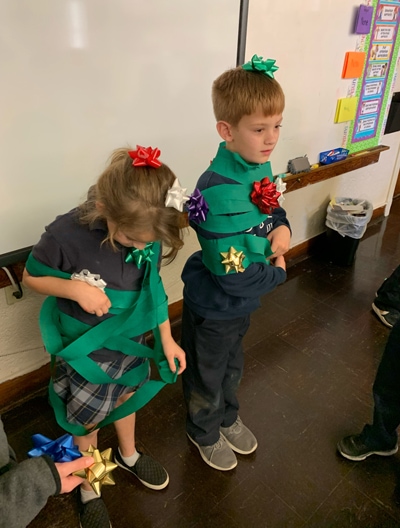 2 elementary school children playing the game 'Human Christmas Tree' with two students wrapped in green streamers and Christmas gift bows to look like a festive Christmas tree.