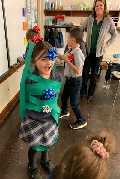 Elementary school children playing the game 'Human Christmas Tree' with one laughing student wrapped in green streamers and Christmas gift bows to look like a festive Christmas tree.